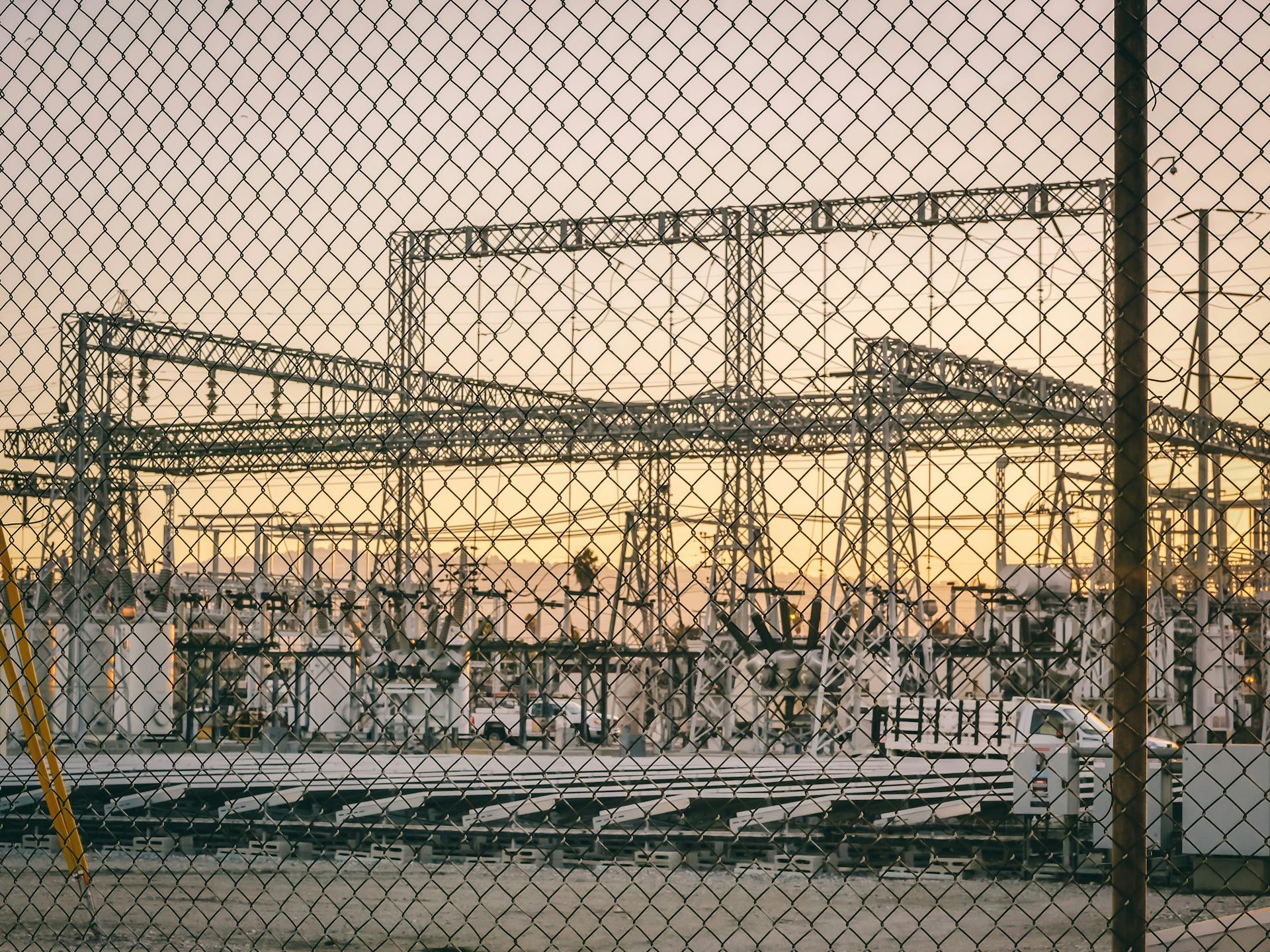 View of an industrial power plant through a chain-link fence at sunrise, symbolizing energy and industry.