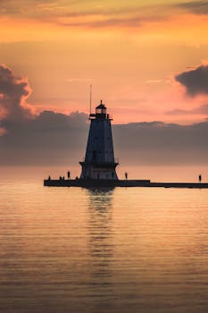 Silhouetted lighthouse with figures against a vibrant sunrise, reflecting on tranquil sea.
