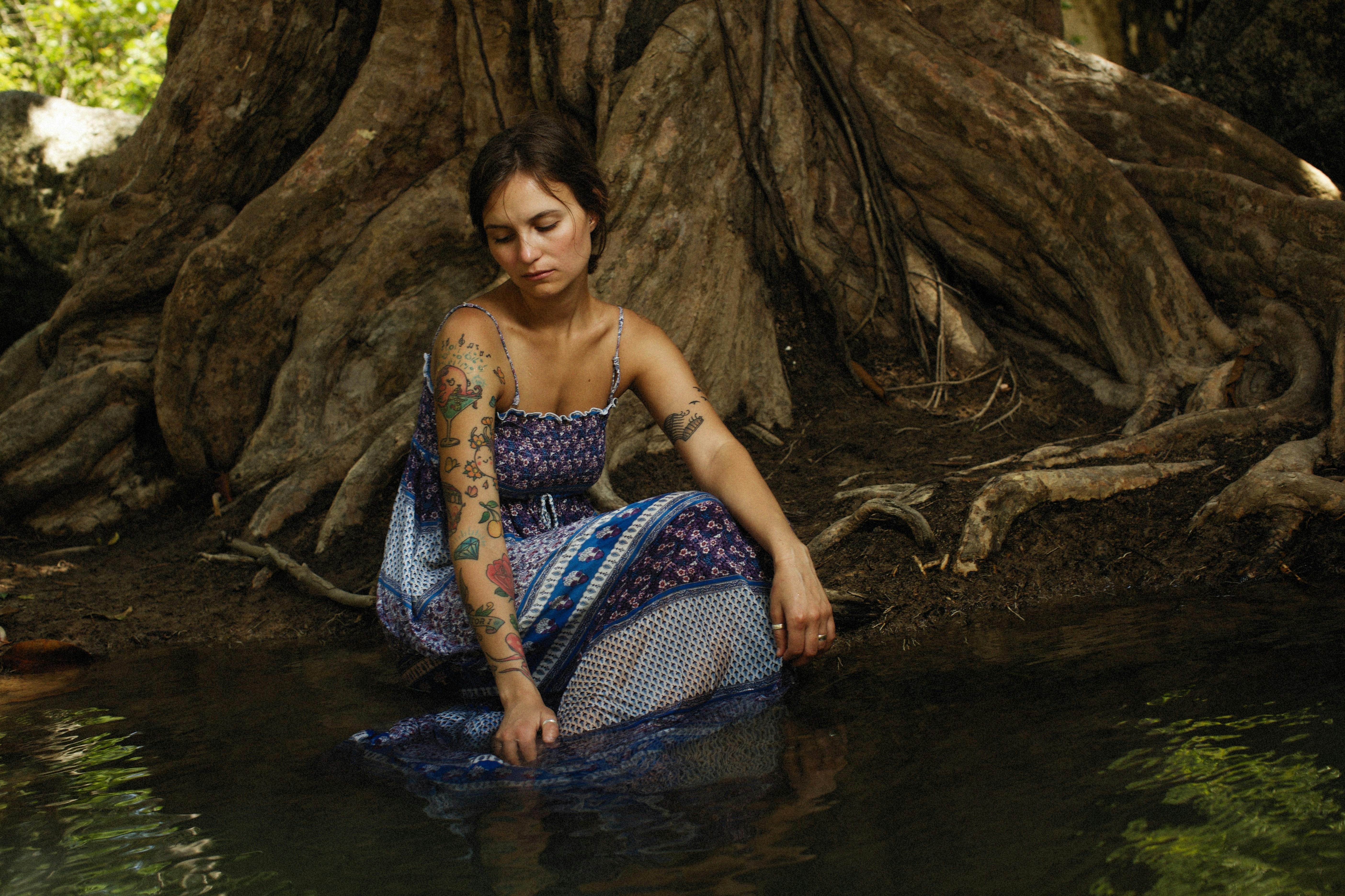 tattooed woman in sleeveless dress sitting in water at foot of big tree