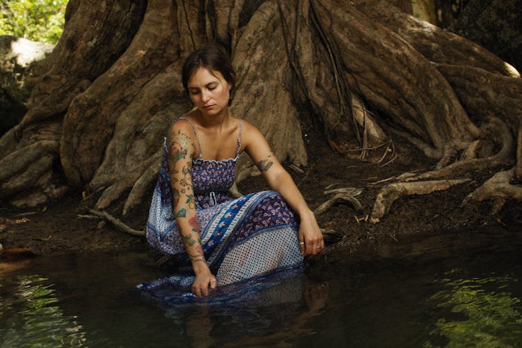 Tattooed Woman In Sleeveless Dress Sitting In Water At Foot Of Big Tree