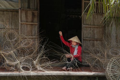 Free An Asian woman in a conical hat crafts wicker baskets outside a rustic wooden building. Stock Photo