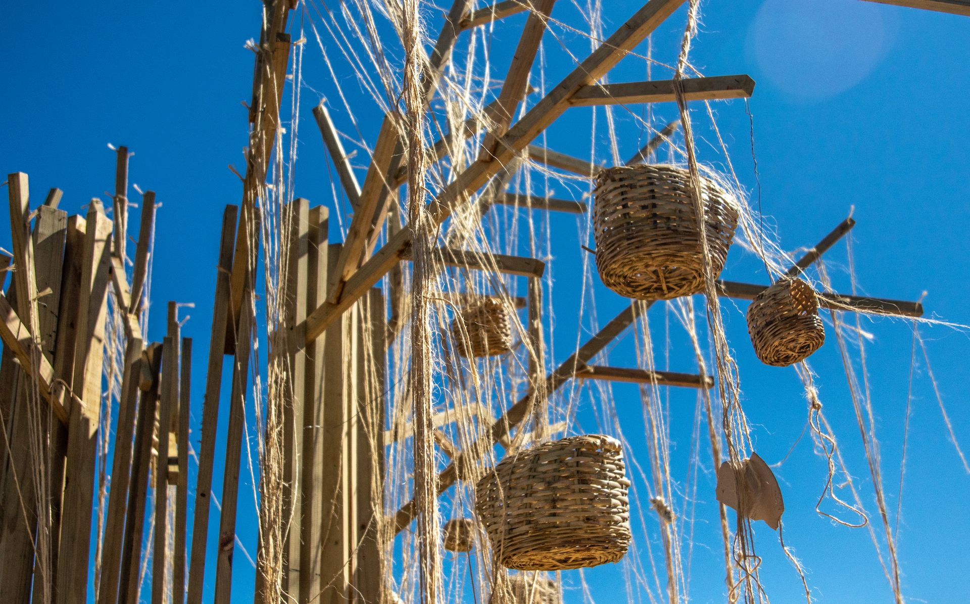 Artistic outdoor display of wicker baskets hanging from wooden structures against a bright blue sky.