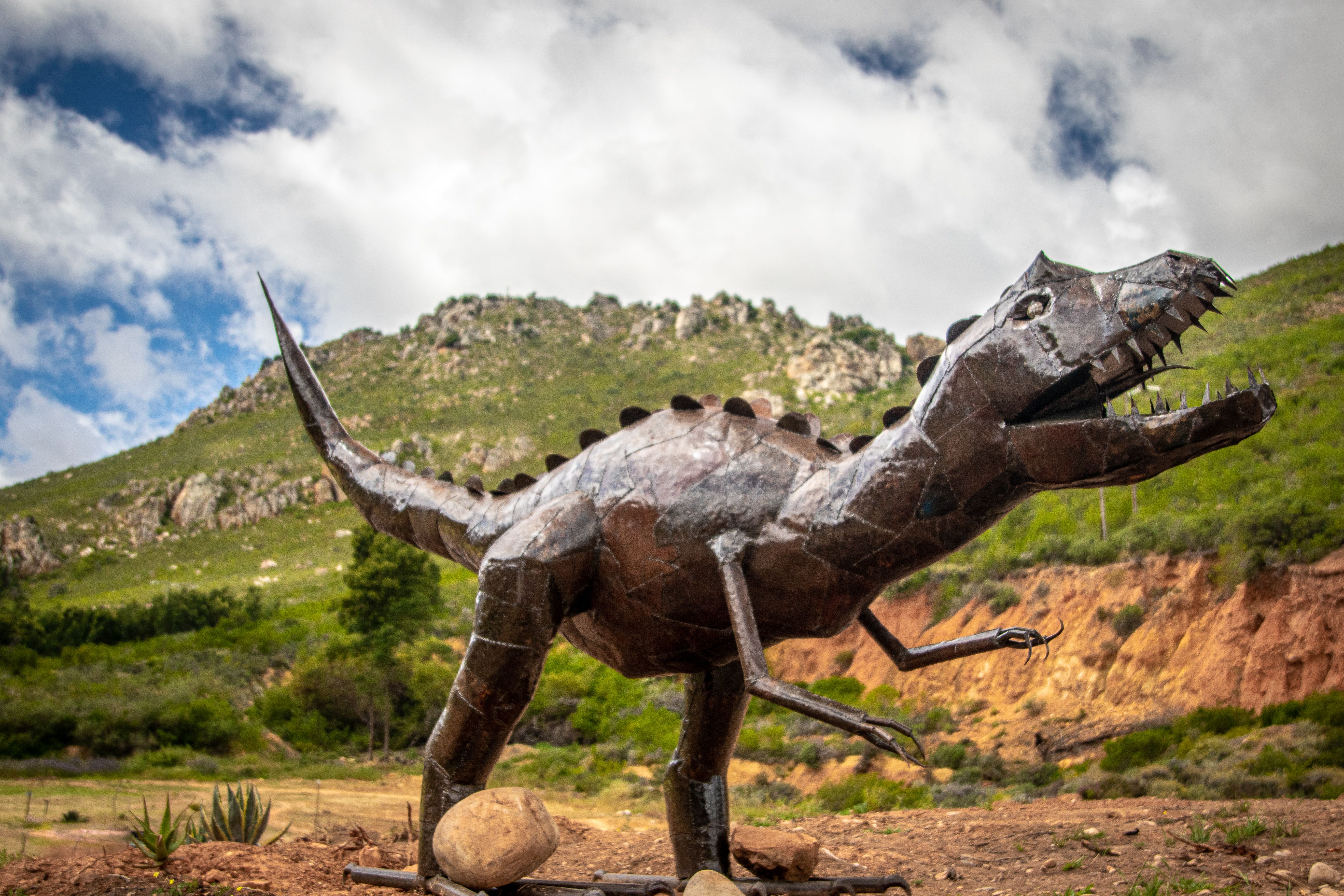A striking metal sculpture of a dinosaur set against a rugged, natural landscape under a cloudy sky.
