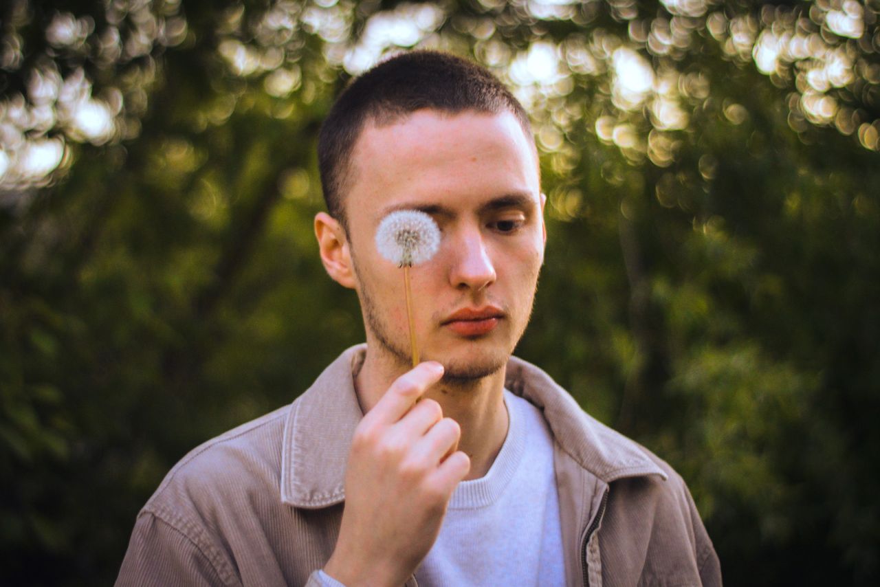 a young man holding a dandelion close to his face in an outdoor settin