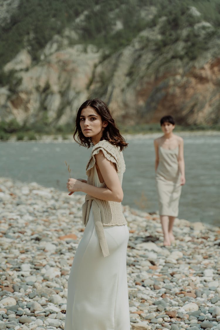 Young Women In Dresses Standing On A Rocky Beach Near A Body Of Water 