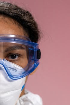 Detailed close-up of a woman wearing safety goggles and a mask against a soft pink background.