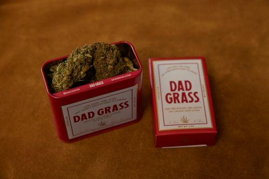 Close-up of a Dad Grass brand tin containing cannabis on a warm surface.