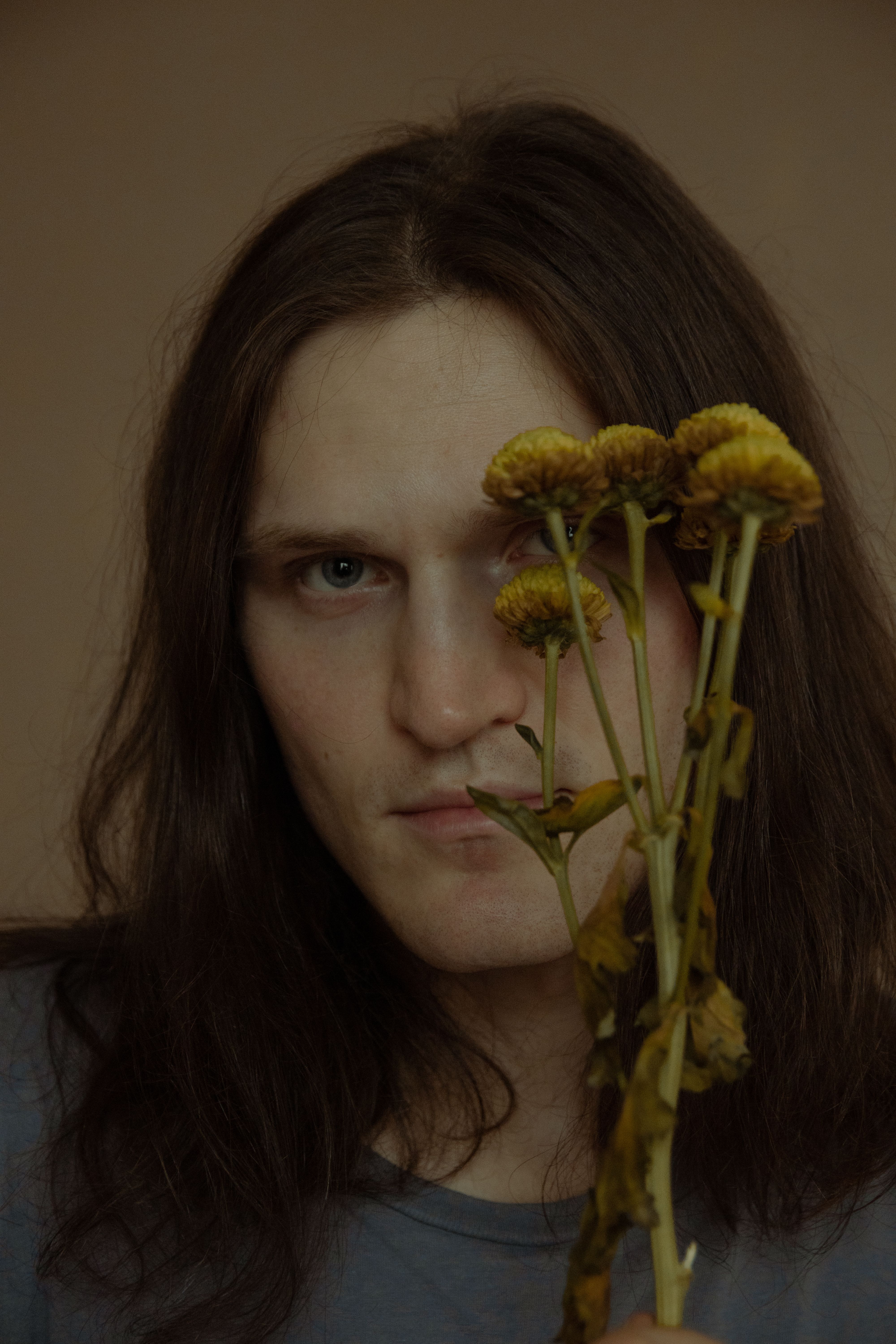 Portrait of a man with long hair holding dried yellow flowers indoors.
