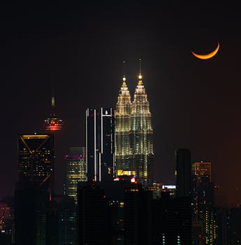 View of Kuala Lumpur skyline featuring Petronas Towers and a crescent moon at night.
