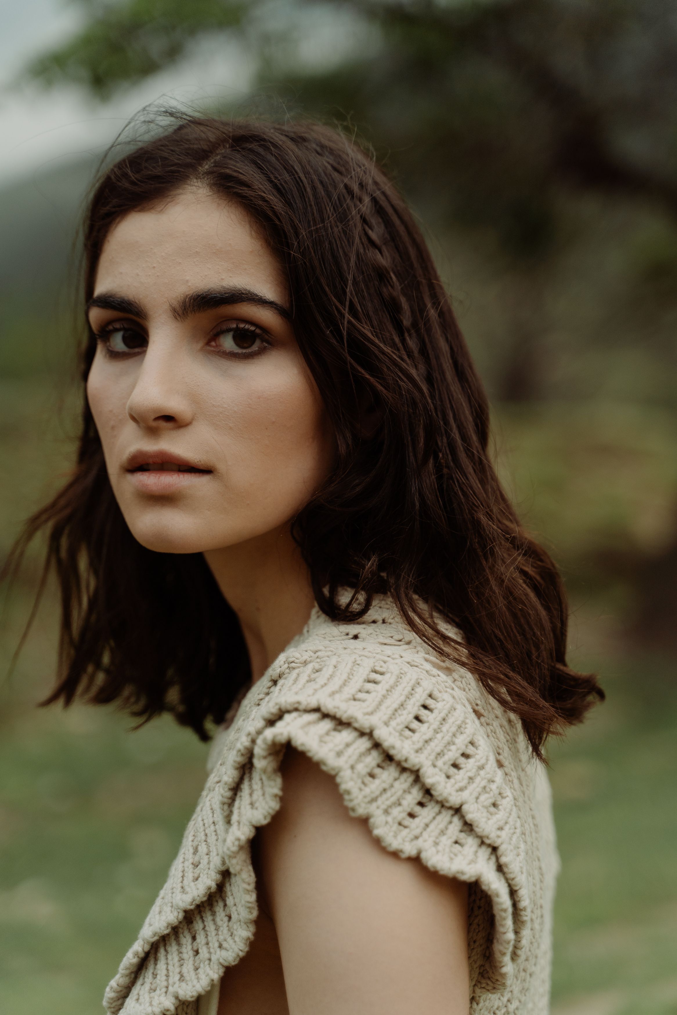 A contemplative woman looking over her shoulder with blurred greenery in the background.