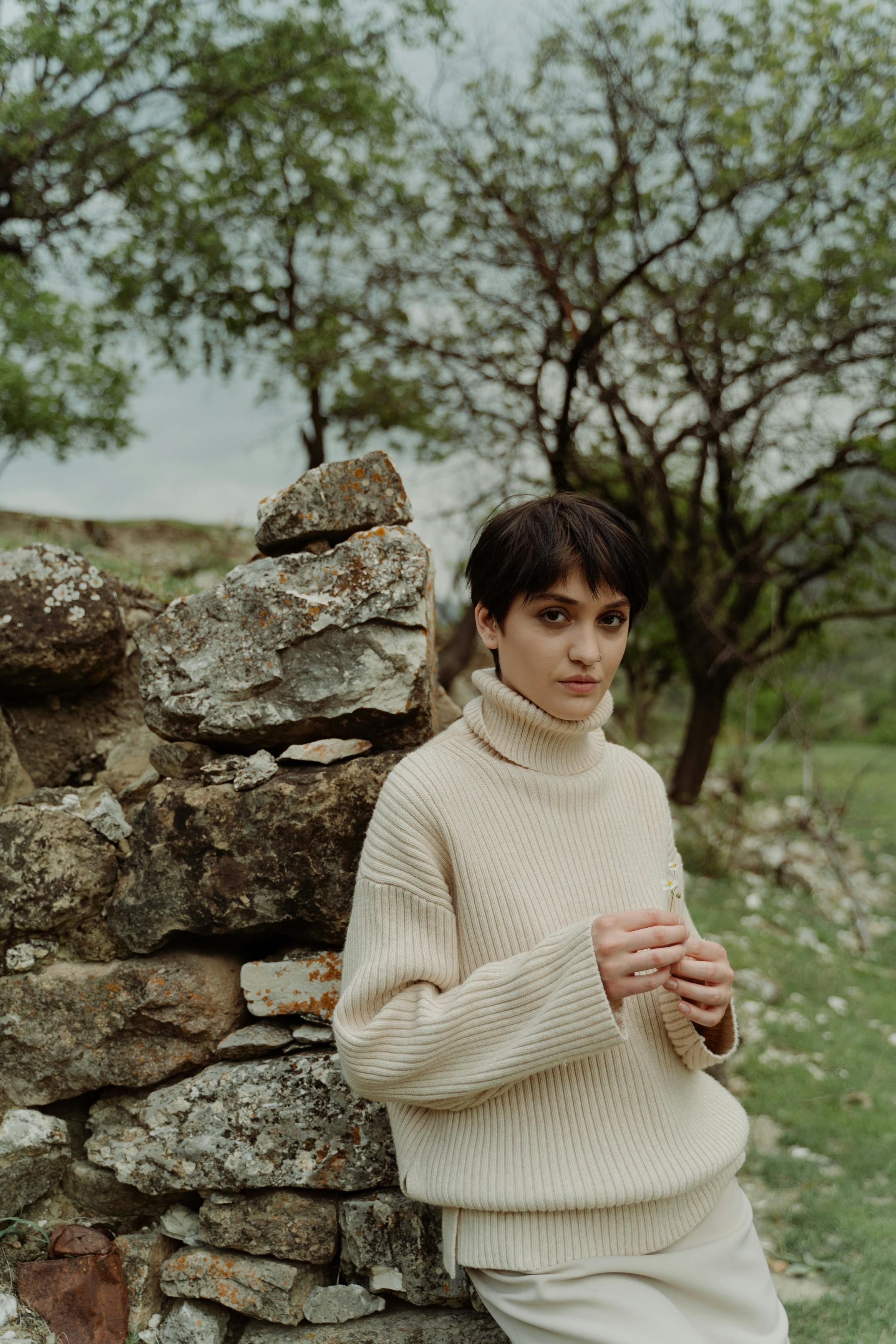 Young woman with short hair in a sweater leaning on a stone wall outdoors with trees in the background.