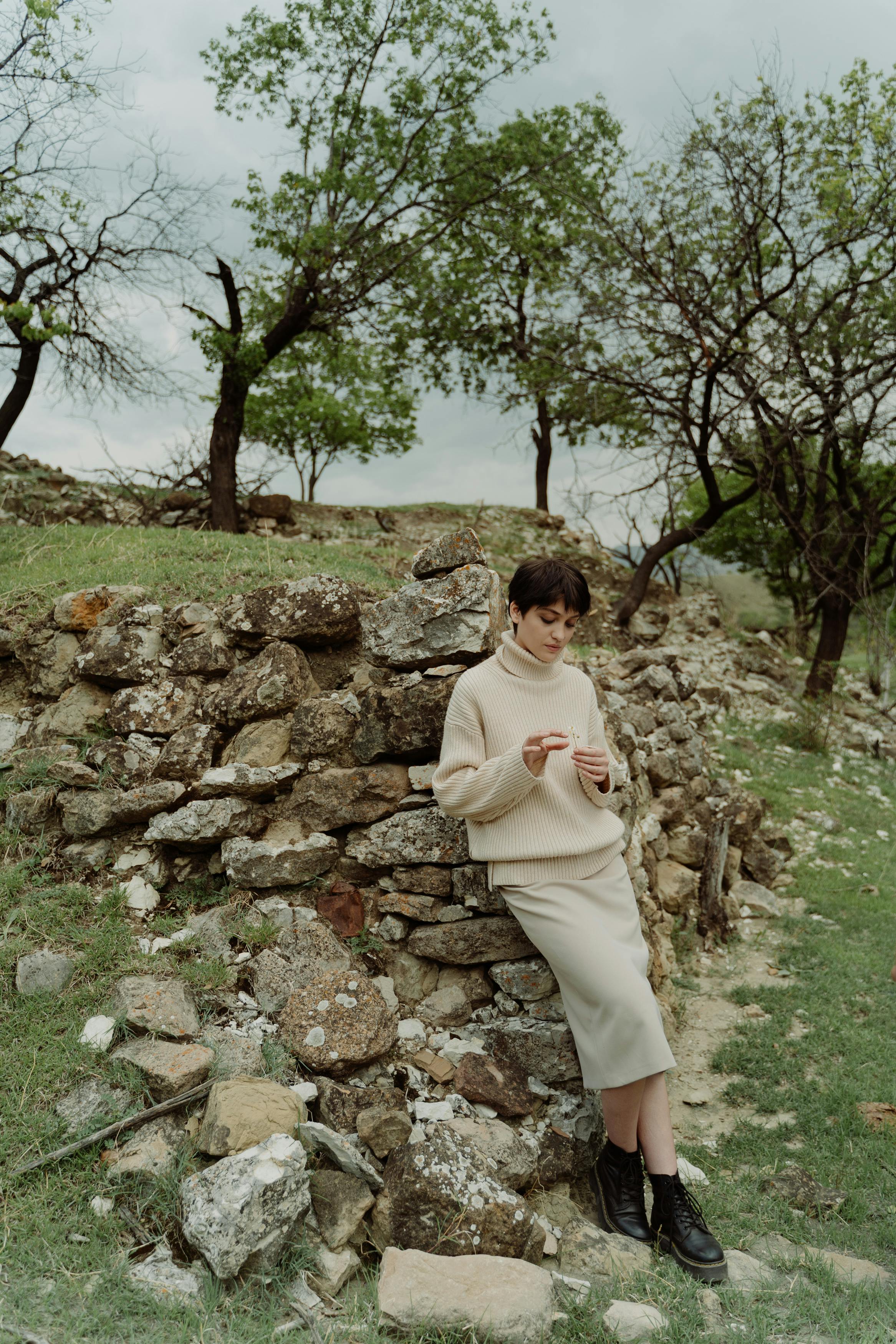 A woman in a cozy sweater leans against a rustic stone wall in a scenic outdoor setting.
