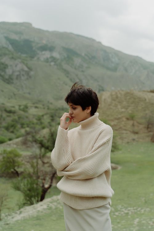 Free A woman in a cozy sweater stands thoughtfully against a serene mountain backdrop. Stock Photo