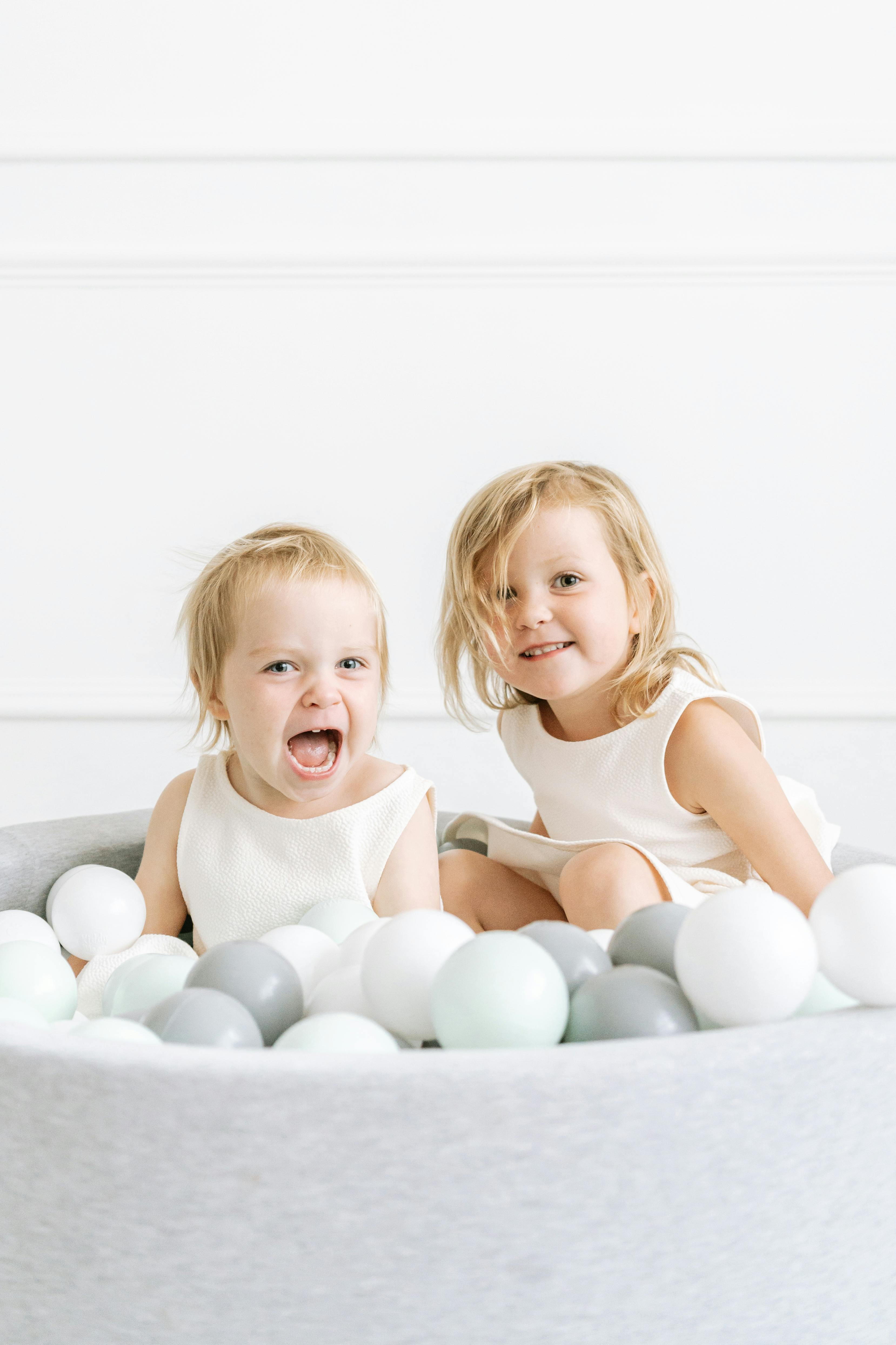 Two smiling children in white dresses enjoy a playful moment in an indoor ball pit.