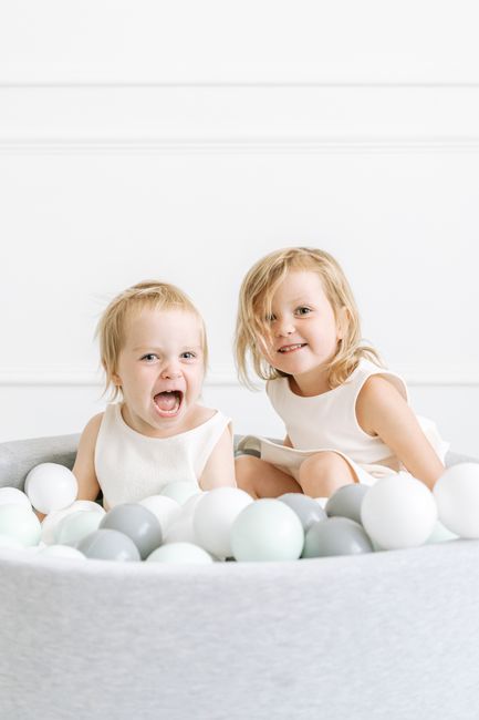Two smiling children in white dresses enjoy a playful moment in an indoor ball pit.