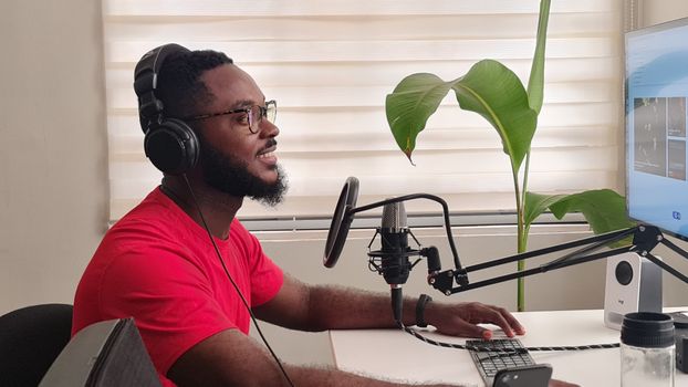 Side view of an African man recording a podcast with microphone and headphones in home office.
