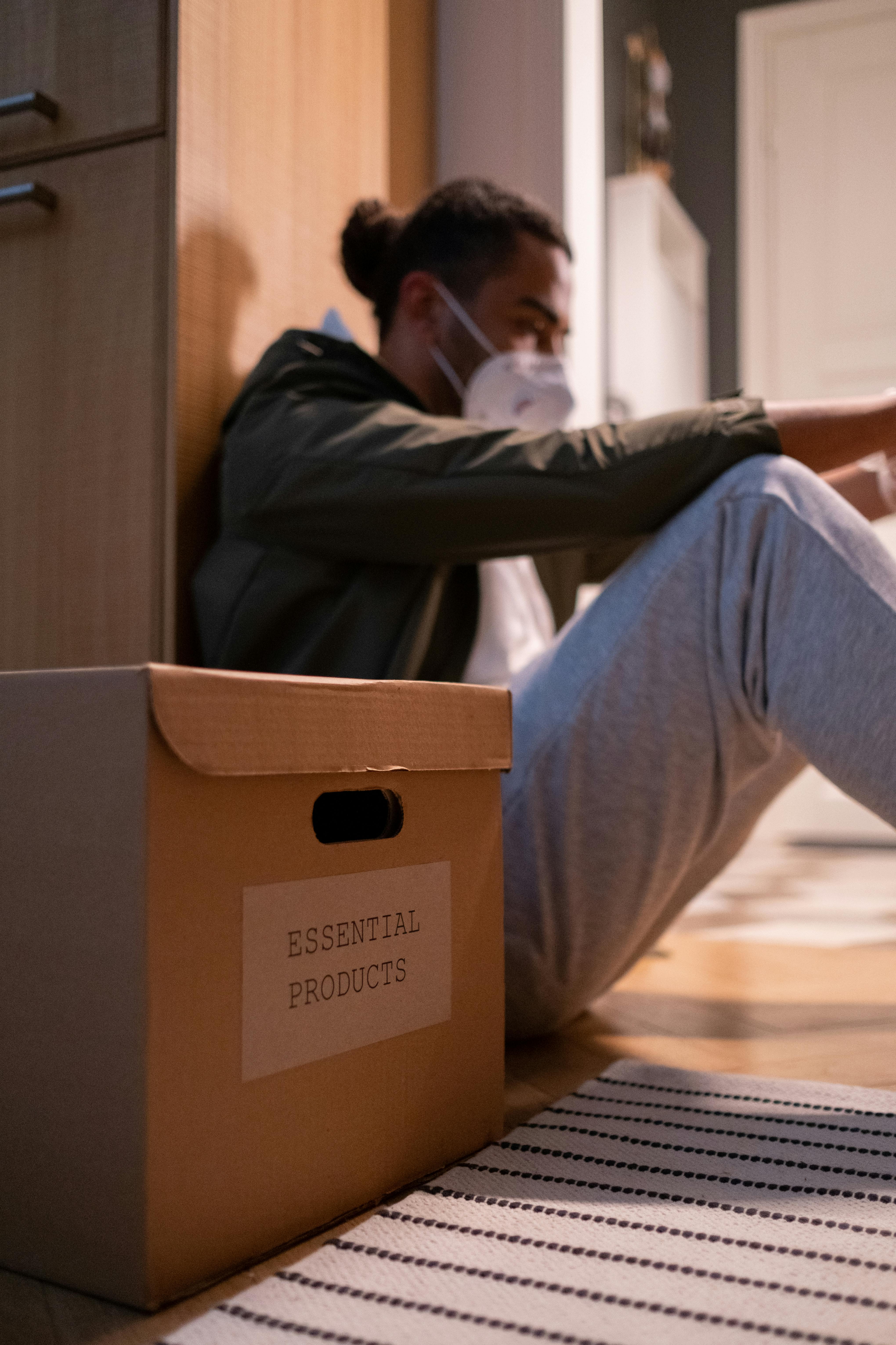 Man wearing a mask sitting by a box labeled 'Essential Products' in a kitchen setting.
