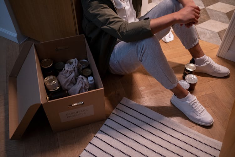 A Person Sitting On The Floor Near A Box Of Canned Food