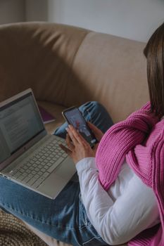 Woman sitting on a sofa multitasking with a smartphone and a laptop indoors.