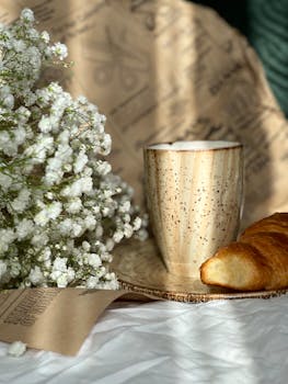 Warm breakfast setting with coffee, croissant, and flowers in sunlight.