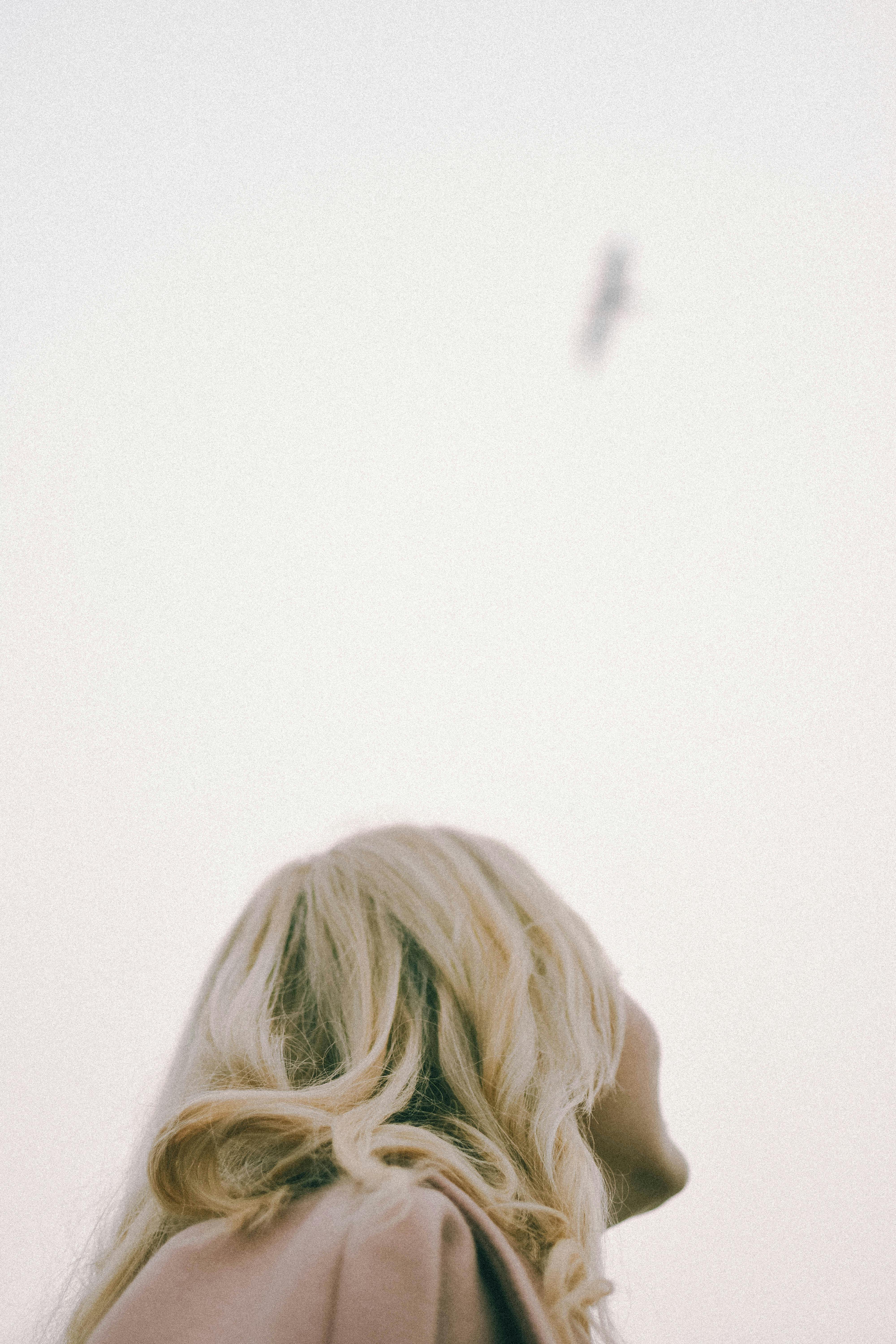 A blonde woman with long hair looks up at a bird flying in an overcast sky.