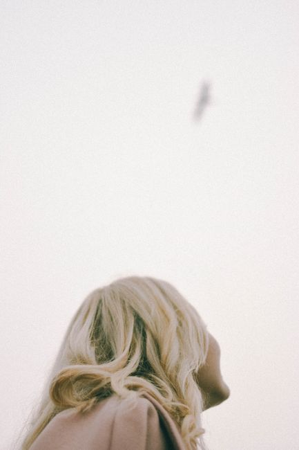 A blonde woman with long hair looks up at a bird flying in an overcast sky.