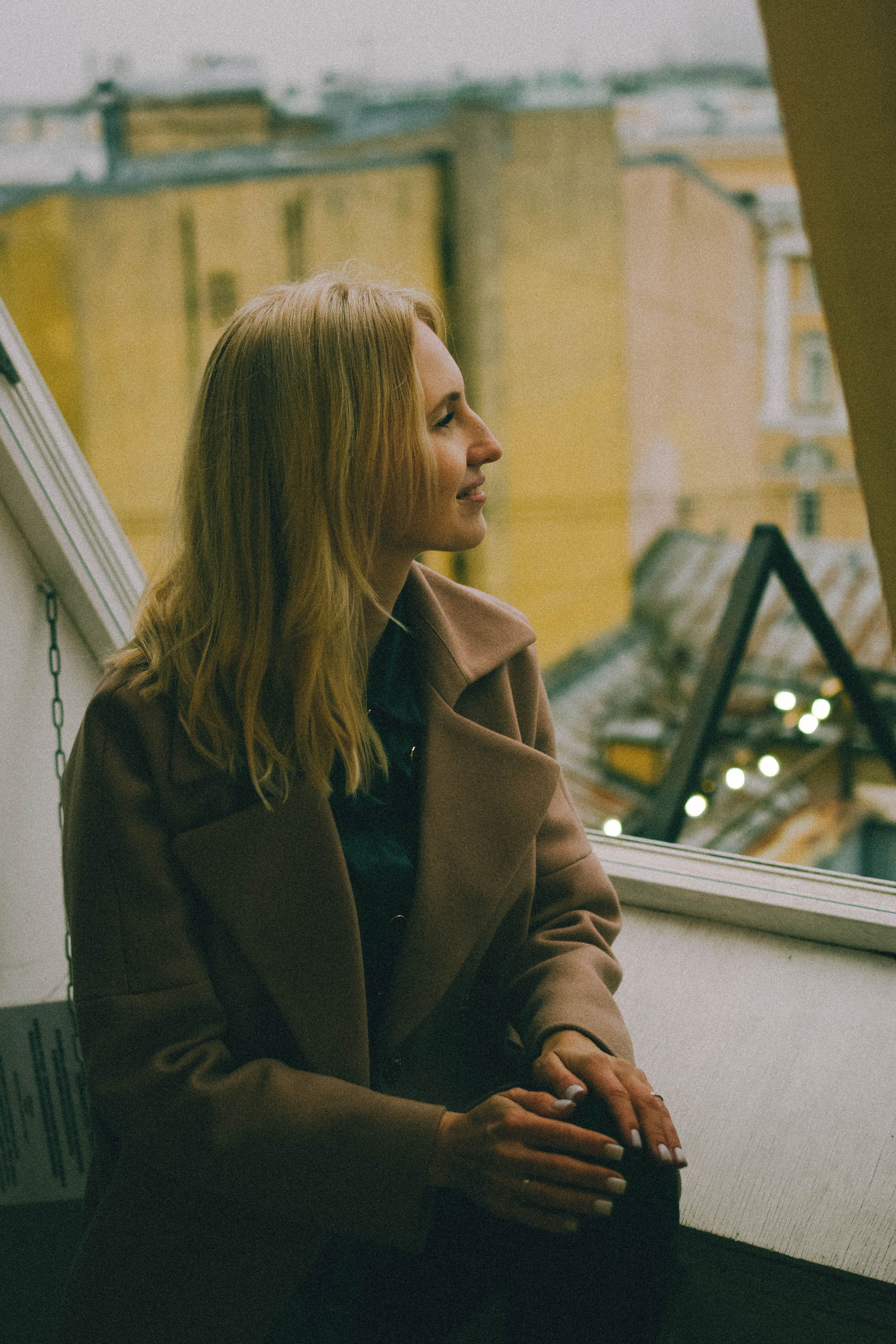 Blonde woman in a coat smiling as she gazes out a window with a blurred cityscape view.