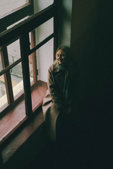 Smiling woman in coat and scarf sits by a window, enjoying a serene indoor moment.