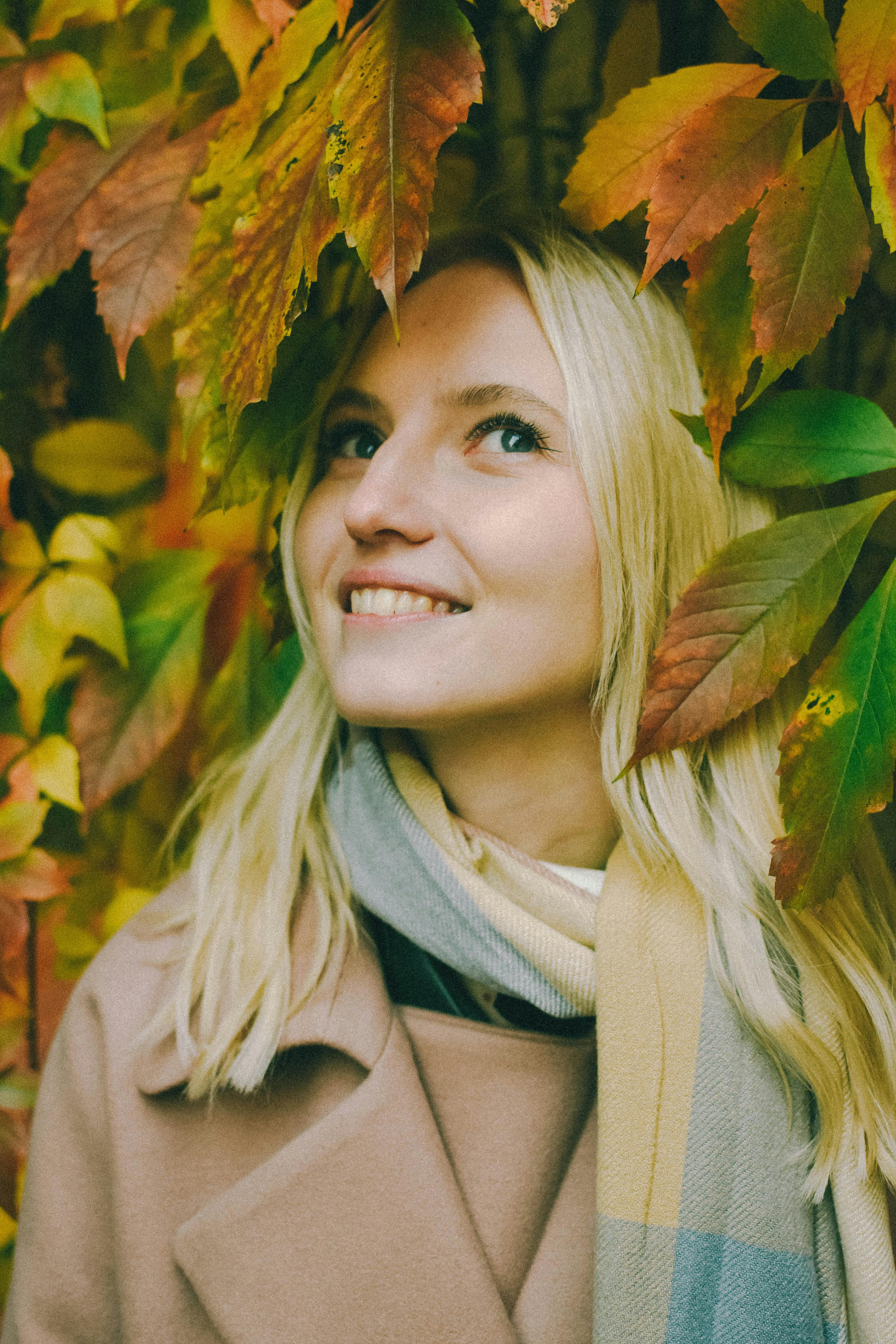 A cheerful woman with blonde hair looks up amidst colorful autumn leaves