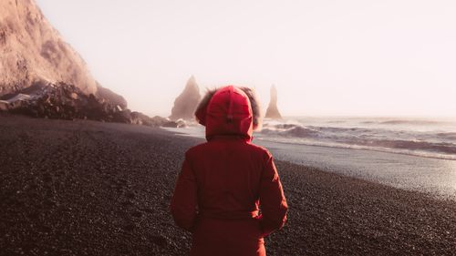 Free A person in a red parka stands on the volcanic sands of Reynisfjara Beach in Iceland, facing sea stacks. Stock Photo