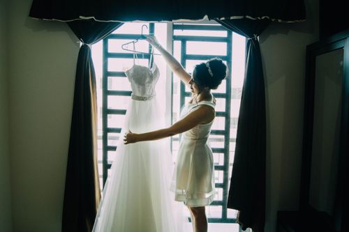 Free A bride in a well-lit room holding her wedding gown in front of a window. Stock Photo