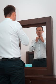 A man in white sleeves adjusts his shirt in front of a mirror indoors, emphasizing reflection and preparation.