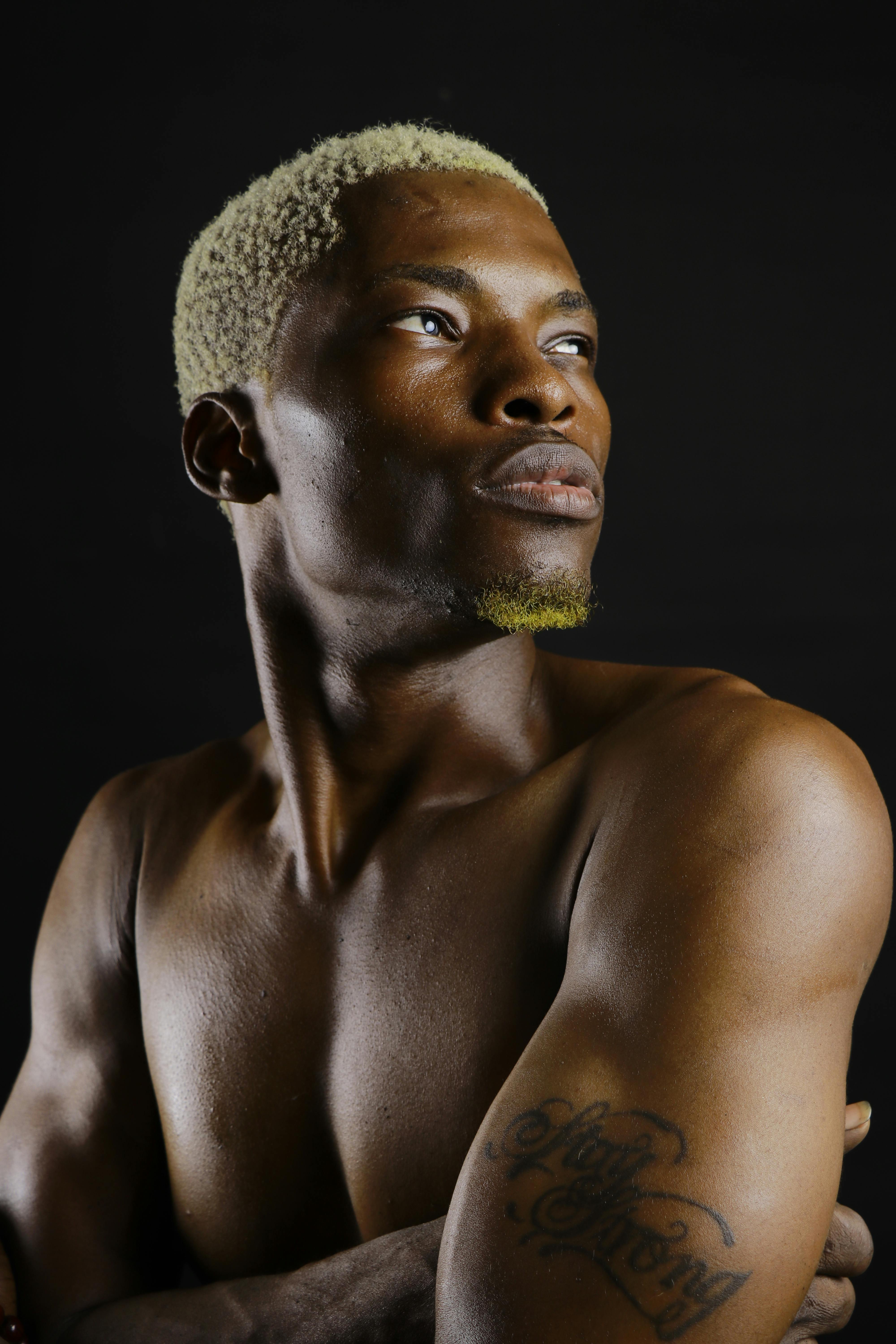 Powerful studio portrait featuring a shirtless muscular man with crossed arms and a facial tattoo.