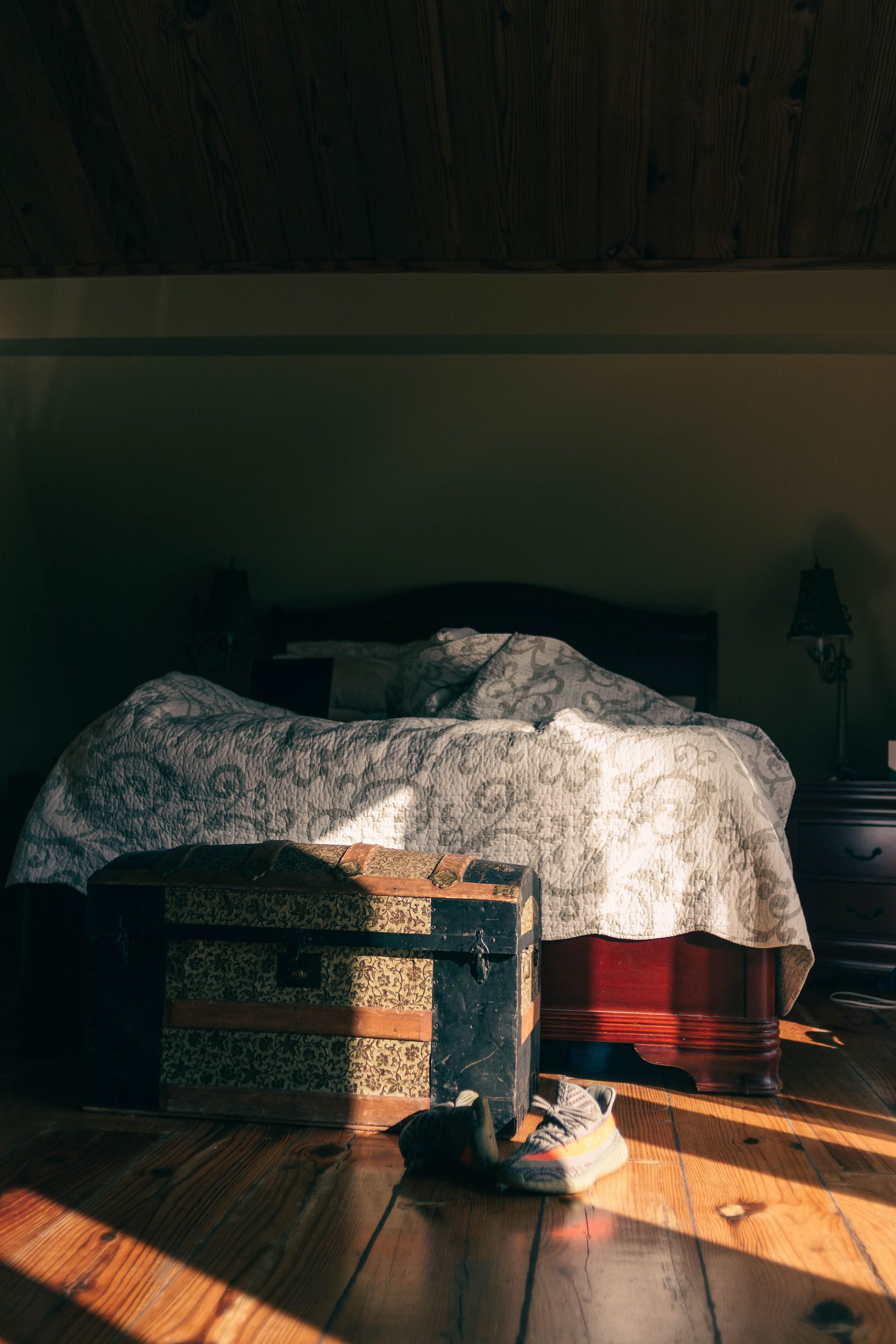 Sunlit bedroom with a bed, old wooden chest, shoes and warm timber flooring