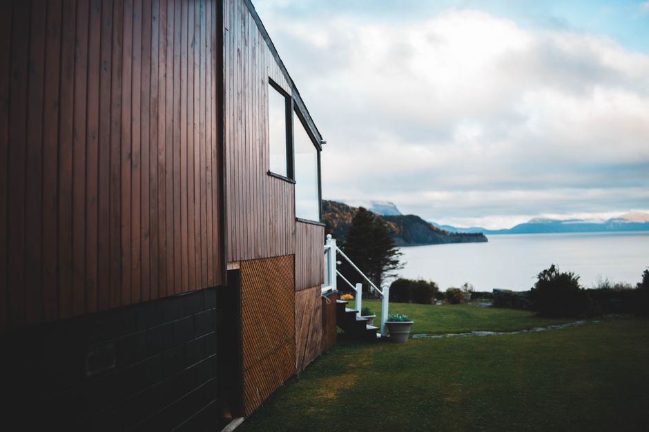 Modernes Ferienhaus mit dunkler Holzfassade an einem Hang mit Blick auf einen Fjord und bewölkten Himmel