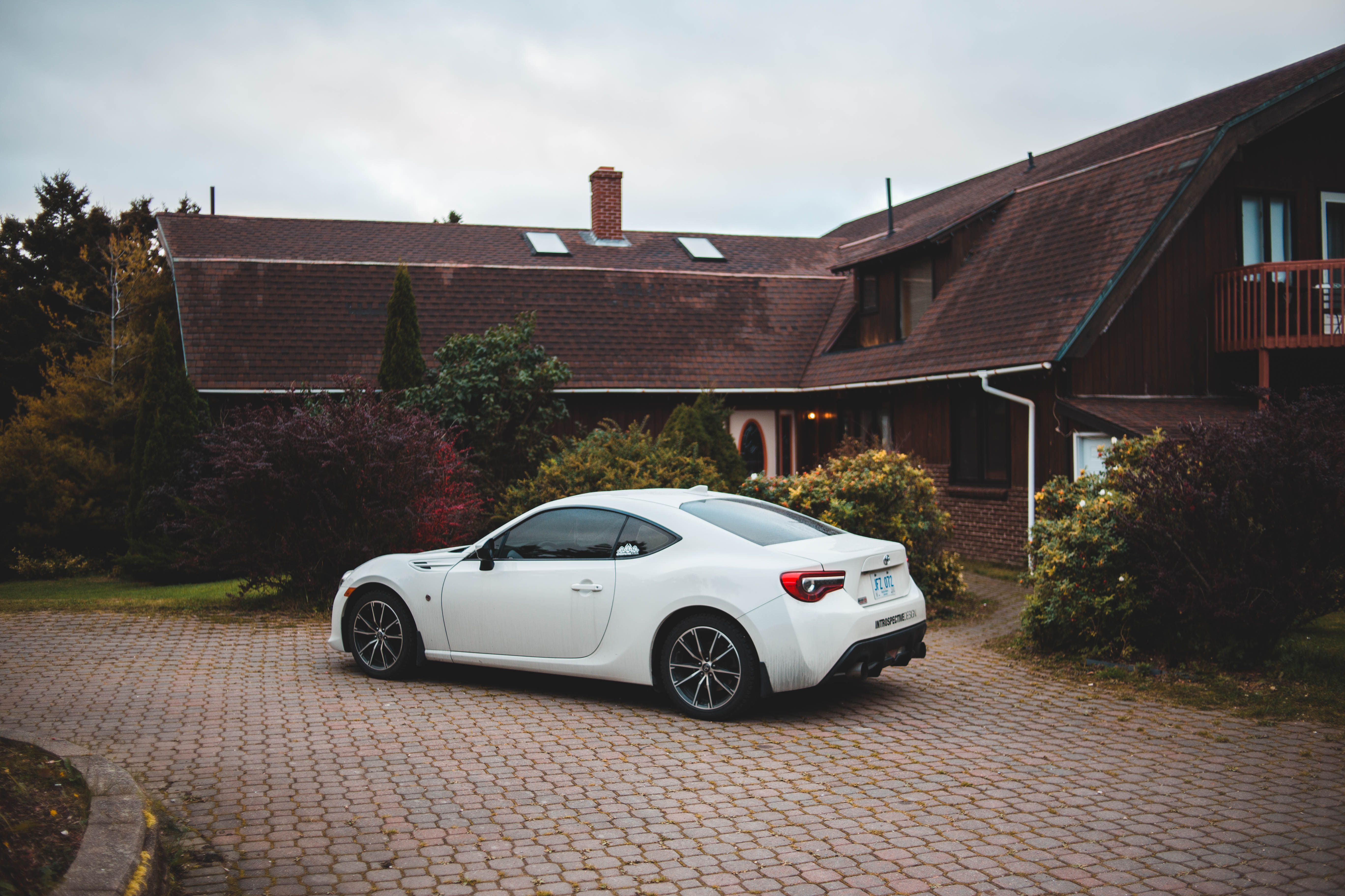 Pflasterarbeiten / Einfahrten / Wege – A sleek white sports car parked in front of a charming rustic house with well-kept gardens.