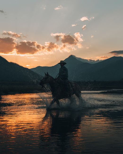 A cowboy rides a horse through a river at sunset in Waterton Park, Alberta, Canada.