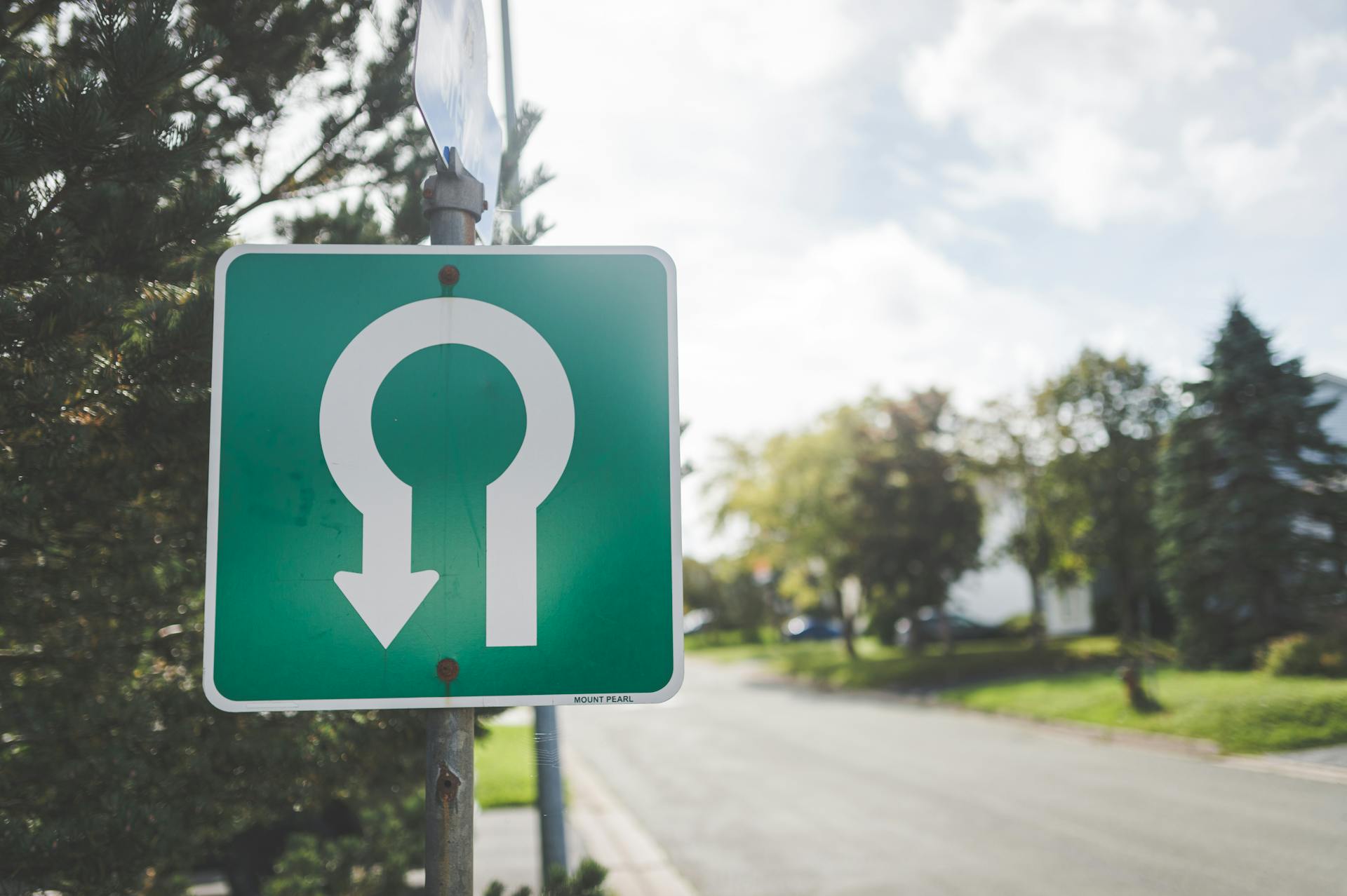 Close-up of a green U-turn road sign on a sunny suburban street with trees.