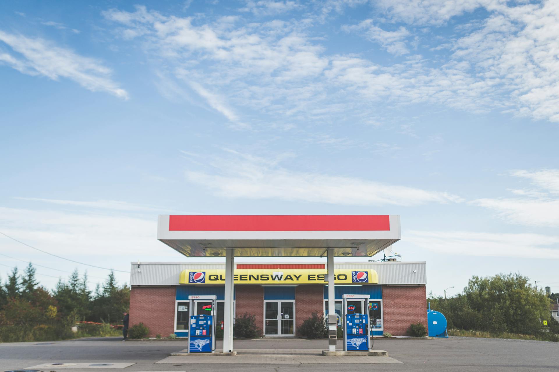 A small, empty gas station with a clear blue sky, featuring a Queensway Esso branding and two fuel pumps.