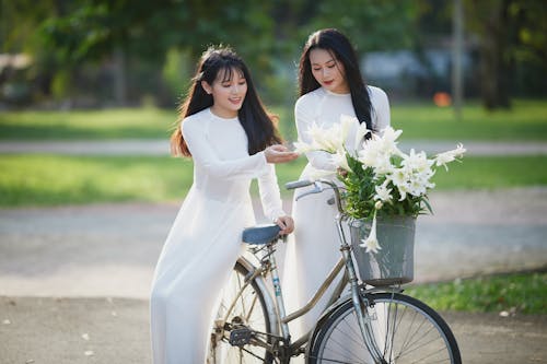 Two Women in White Dresses Looking at Flowers in Bicycle Basket