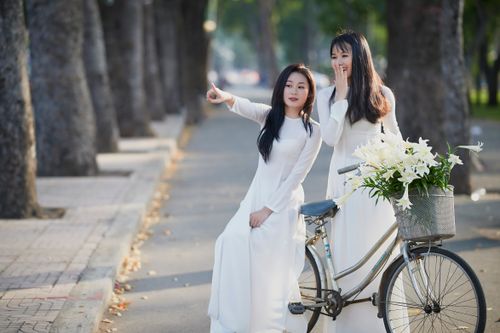 Free Two women in white dresses with a bicycle adorned with lilies, enjoying a sunny day outdoors. Stock Photo