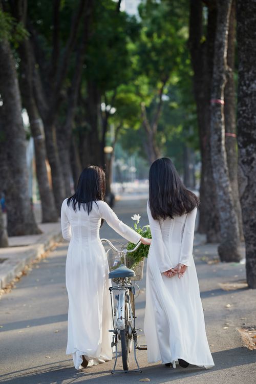 Free Two women in elegant white dresses walk a bicycle down a tree-lined path, embodying serene companionship. Stock Photo