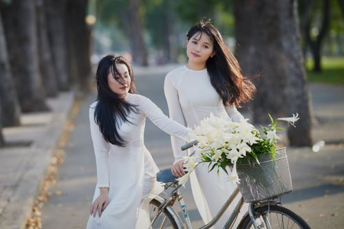 Free Two Asian women in white dresses with a bicycle in a sunlit park. Elegant and serene vibes. Stock Photo