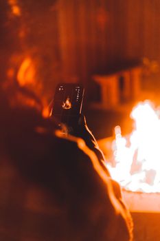 A person photographs a bonfire with a smartphone, surrounded by warm firelight.