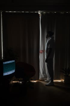A solitary man in a white shirt stands by dark curtains, illuminated softly, with a hint of mystery.
