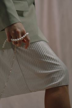Close-up of a woman in a green blazer holding a string of pearls, wearing a pleated skirt.
