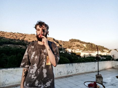 Stylish man standing on a rooftop against a scenic hillside backdrop, under the clear sky.