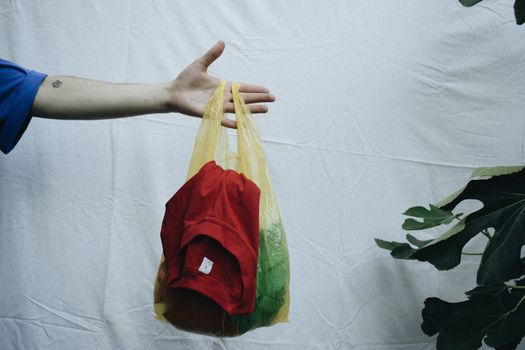 A hand holds a yellow plastic bag containing clothes on a simple white background.
