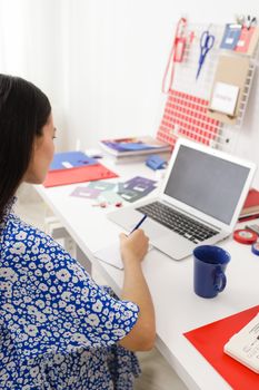 A woman in a blue dress works on a laptop in a bright, organized home office setup.