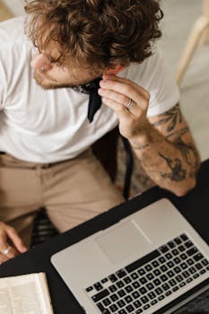 A tattooed man adjusts his ear with a laptop in view, creating a modern and stylish workspace feel.